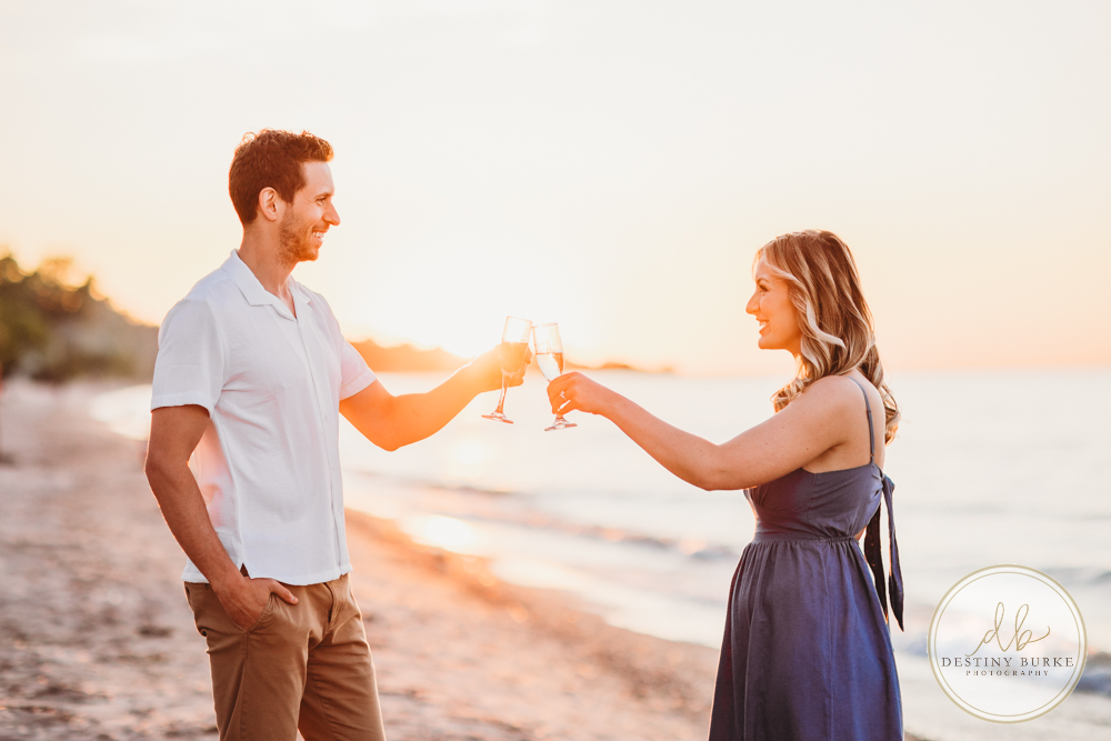 Engagement Session with Couple at Durand Eastman Beach in Irondequoit, NY near Rochester at sunset with champagne toast, engagement ring